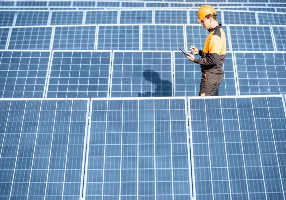 View on the rooftop solar power plant with engineer in protective workwear walking and examining photovoltaic panels. Concept of alternative energy and its maintenance